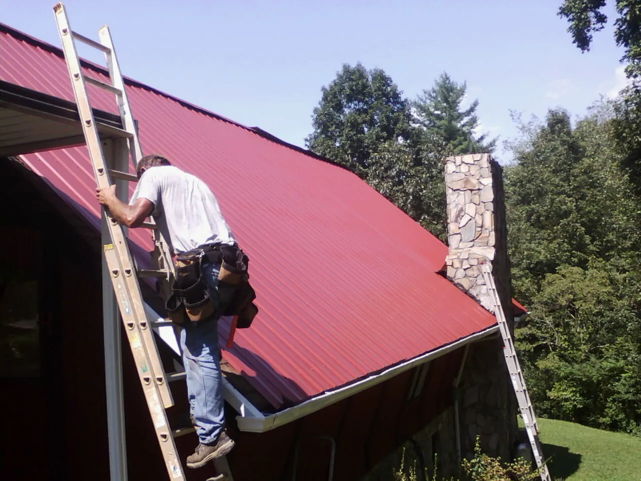 Roofer on a ladder beside a red metal roof and stone chimney — Soffit & Fascia Repair by Roofing Brothers in Camak
